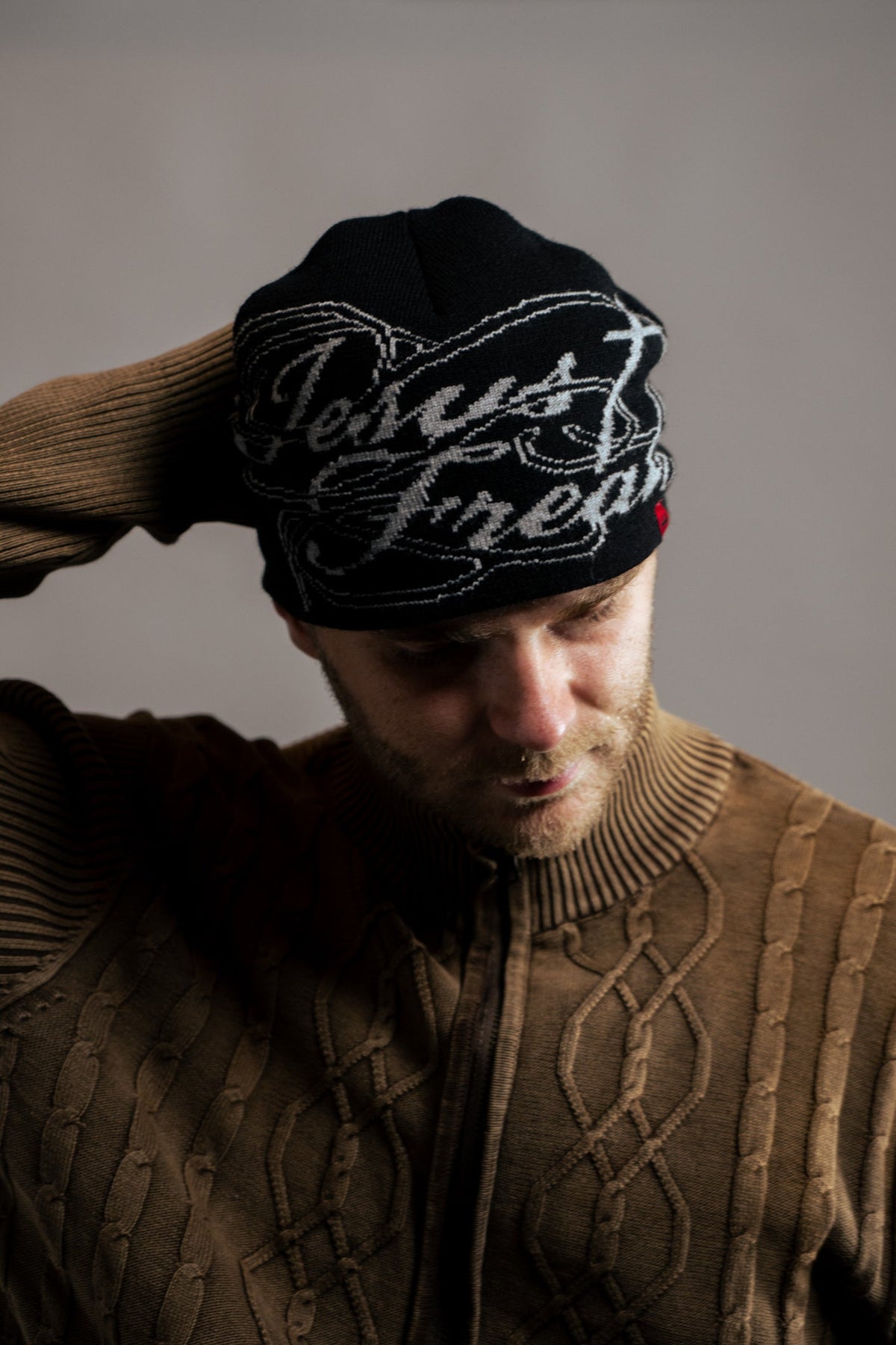 A man wearing a black knit beanie with white cursive embroidery that reads 'Jesus Fnepp' and a brown cable-knit sweater, looking down in a dimly lit setting.